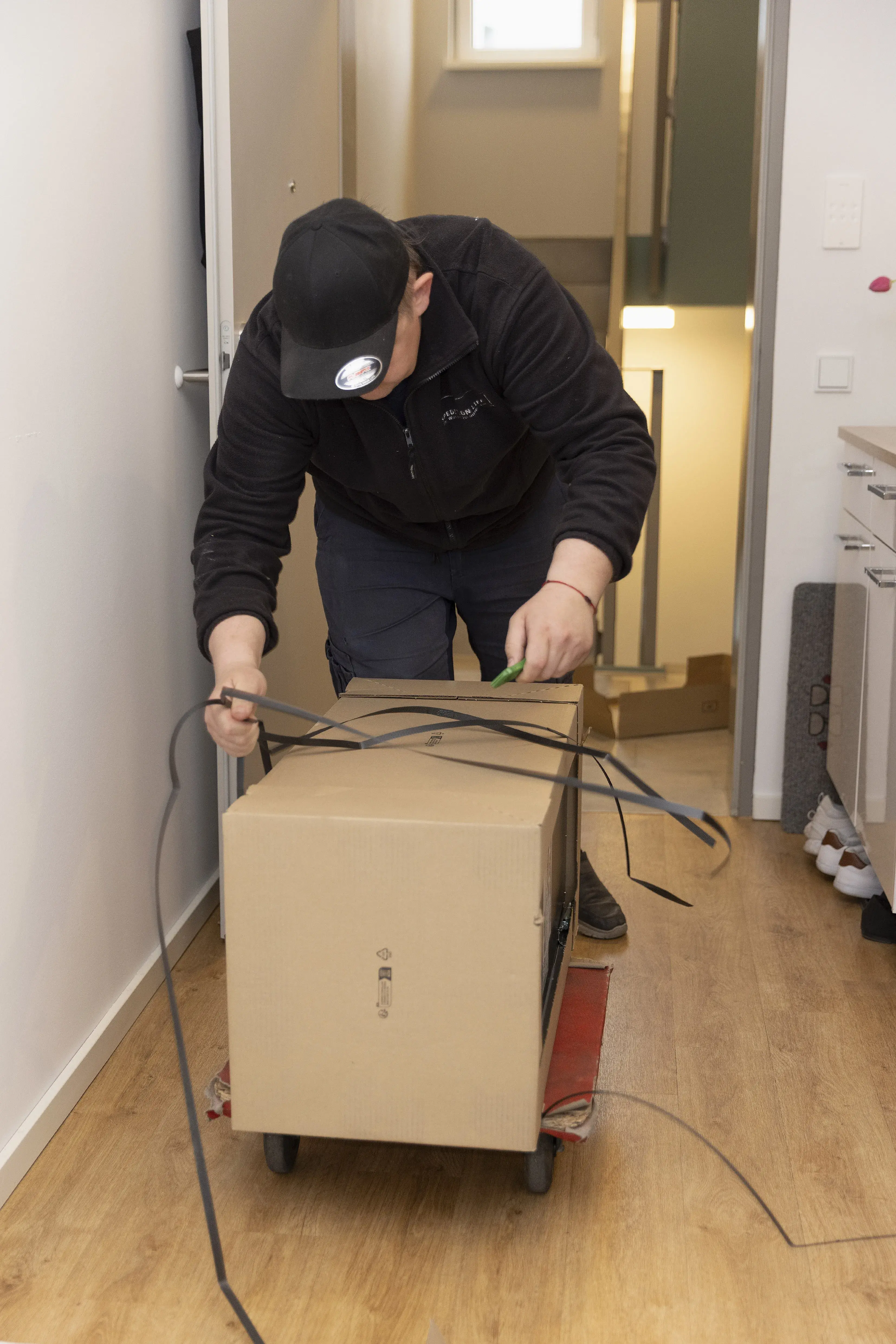 Mann bindet Kabel um einen Karton auf einem Rollwagen in einem Flur.